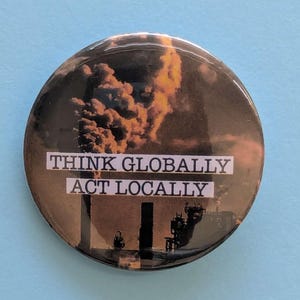 May include: A round button with a black background and a picture of a factory with smoke coming out of the chimney. The text "THINK GLOBALLY ACT LOCALLY" is printed in white on a black rectangle.