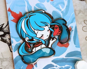 May include: A blue and white enamel pin featuring a cartoon character with blue hair holding a red fish. The pin is on a white card with blue and red watercolor-like designs. The card also has the text "ENAMEL PIN" and "HIDEAWAYMELON.COM".