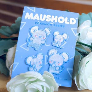 May include: A light blue rectangular backing card featuring the text MAUSHOLD POKEMON SERIES in white, displaying four embroidered patches of white mouse-like characters with pink cheeks and blue outlines, arranged in a grid pattern on the card.
