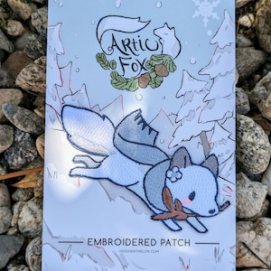 Könnte beinhalten: Gestickter Aufnäher mit einem blauen Fuchs, der durch einen schneebedeckten Wald rennt. Der Fuchs hält einen Ast im Maul und trägt eine Blume auf dem Kopf. Der Aufnäher ist mit "Embroidered Patch" und "Hideawaymelon.com" beschriftet.