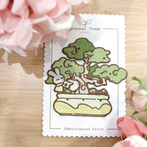 Pode incluir: Etiqueta bordada com uma árvore bonsai com um pequeno coelho sentado no vaso. A etiqueta está em um cartão branco com o texto "Bonsai Tree Series" e "Embroidered Patch".