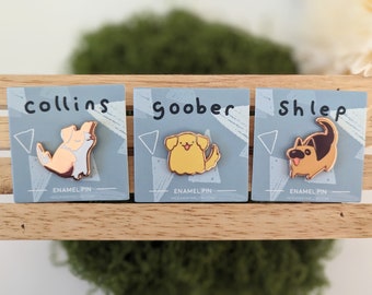 May include: Three enamel pins featuring cartoon dogs. The pins are on a wooden surface. The pins are labeled "Collins", "Goober", and "Shlep".