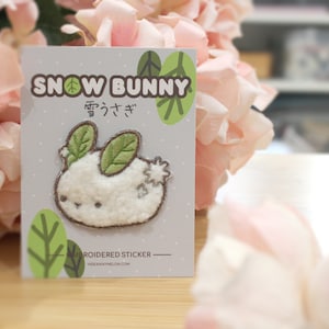 Op de afbeelding: Een witte, pluche, geborduurde konijnen sticker met groene bladeren als oren. De sticker zit op een kaart met de tekst "SNOW BUNNY" en Japanse karakters. De kaart heeft een groen bladontwerp.
