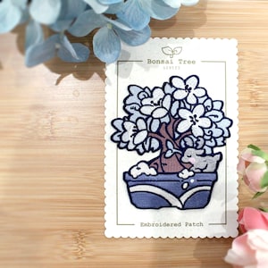 May include: Embroidered patch featuring a blue bonsai tree with white flowers and a small gray animal sitting in the pot. The patch is on a white card with the text "Bonsai Tree Series" and "Embroidered Patch".