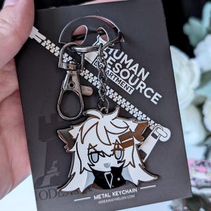 May include: A metal keychain featuring a cartoon character with white and brown hair, a black outfit, and gray eyes. The keychain is attached to a black card with the text "HUMAN RESOURCE DEPARTMENT" and "METAL KEYCHAIN".