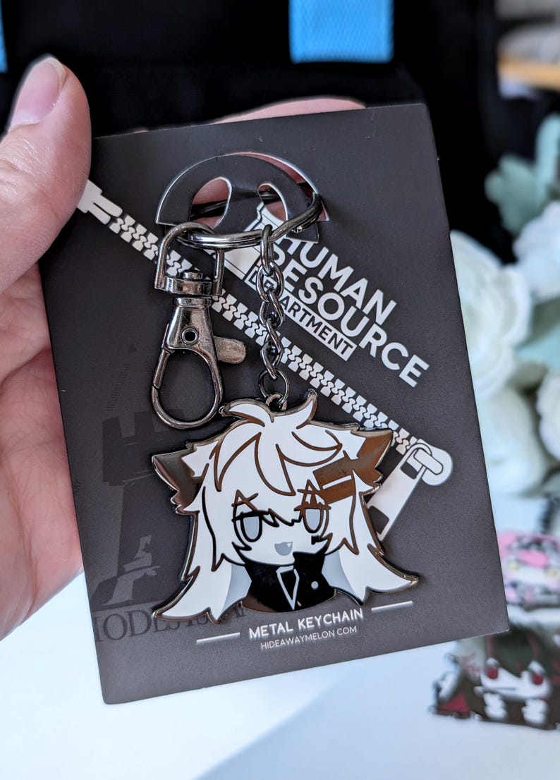 May include: A metal keychain featuring a cartoon character with white and brown hair, a black outfit, and gray eyes. The keychain is attached to a black card with the text "HUMAN RESOURCE DEPARTMENT" and "METAL KEYCHAIN".