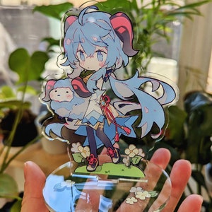 May include: A blue and pink anime character with long blue hair and red horns holding a small white fluffy animal. The character is standing on a clear acrylic base with white flowers.