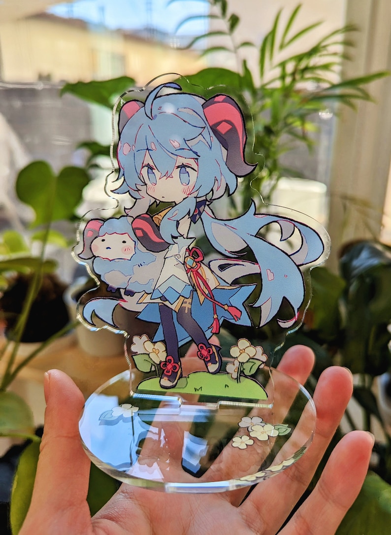 May include: A blue and pink anime character with long blue hair and red horns holding a small white fluffy animal. The character is standing on a clear acrylic base with white flowers.