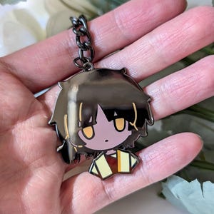 May include: A black metal keychain featuring a cartoon character with black hair and yellow eyes. The character is wearing a yellow and brown outfit. The keychain is attached to a black chain.