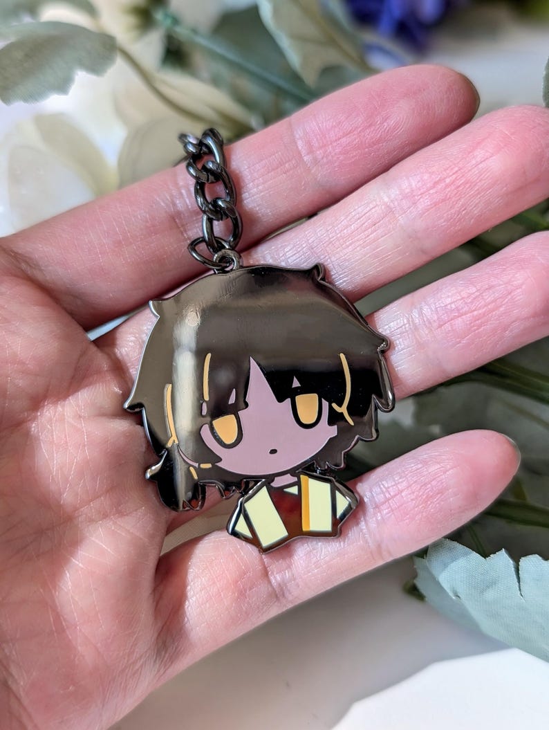 May include: A black metal keychain featuring a cartoon character with black hair and yellow eyes. The character is wearing a yellow and brown outfit. The keychain is attached to a black chain.
