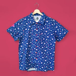 Puede incluir: Una camisa azul de manga corta con un estampado de huesos blancos, corazones rosas y cerebros rosas. La camisa tiene botones rojos y un pequeño bolsillo. La camisa está colgada de una percha de madera sobre un fondo rojo.
