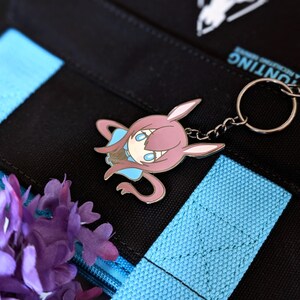 May include: A pink and blue cartoon character keychain with a silver keyring. The keychain is attached to a black bag with blue straps. Purple flowers are in the lower left corner. The bag has white text and a white logo.
