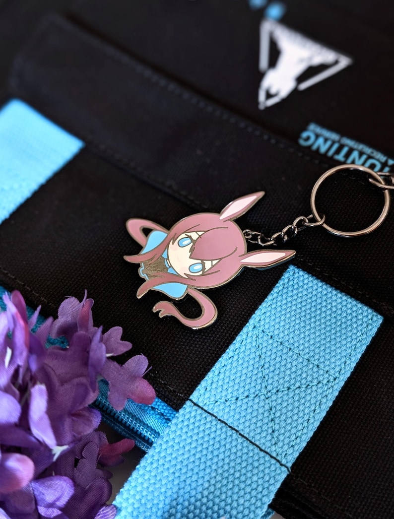 May include: A pink and blue cartoon character keychain with a silver keyring. The keychain is attached to a black bag with blue straps. Purple flowers are in the lower left corner. The bag has white text and a white logo.