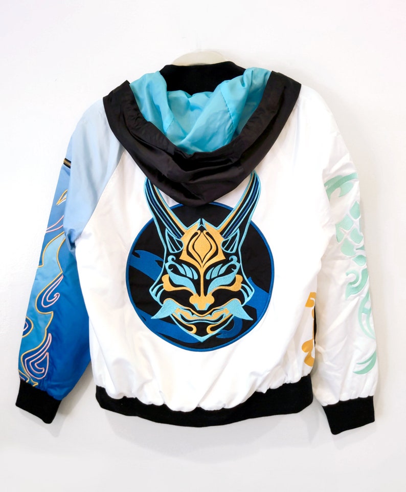 May include: White hooded jacket with a blue and gold mask design on the back. The mask is in a circle with blue and white waves around it. The jacket has blue and white sleeves with a gold and white design.
