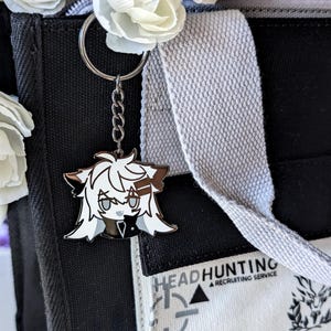 May include: A silver keychain with a cartoon character charm. The charm features a white and brown anime-style character with a black outfit. The keychain is attached to a black tote bag with the text "HEADHUNTING A RECRUITING SERVICE". White flowers are in the bag.