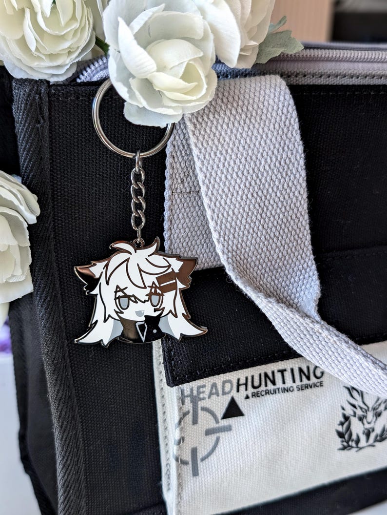 May include: A silver keychain with a cartoon character charm. The charm features a white and brown anime-style character with a black outfit. The keychain is attached to a black tote bag with the text "HEADHUNTING A RECRUITING SERVICE". White flowers are in the bag.