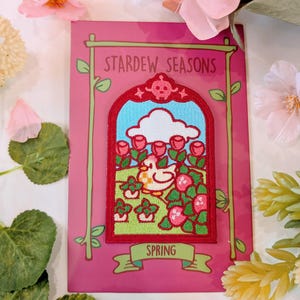 Puede incluir: Una tarjeta rosa con un parche bordado rojo que representa a un pollo de dibujos animados en un jardín de primavera. El parche está enmarcado con un borde dorado y el texto "Stardew Seasons" y "Spring" está impreso en la tarjeta.