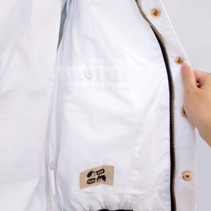 May include: White zippered jacket with a black zipper and gold zipper pulls. The jacket has a small pocket on the inside with a button closure. The jacket has a label on the inside that says "MEOW" and "HYPE".