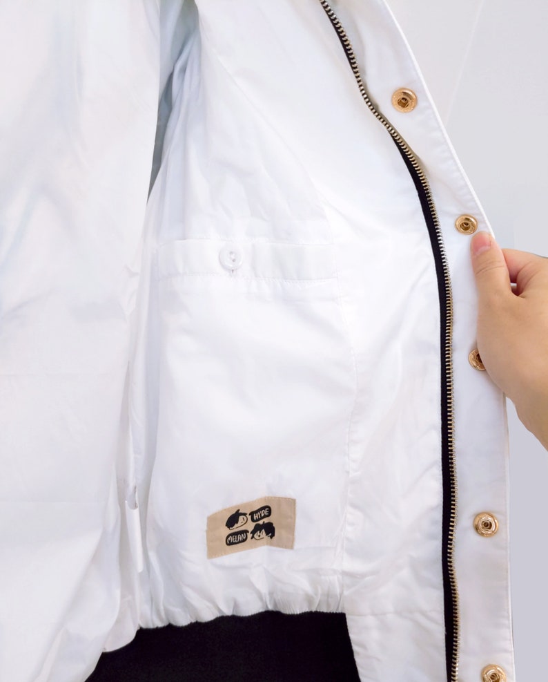 May include: White zippered jacket with a black zipper and gold zipper pulls. The jacket has a small pocket on the inside with a button closure. The jacket has a label on the inside that says "MEOW" and "HYPE".