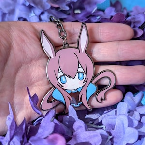 May include: Pink and blue anime-style bunny keychain. The keychain features a cartoon bunny with large blue eyes, pink hair, and a blue collar. The keychain is made of metal and has a black chain. The background is filled with purple flowers.