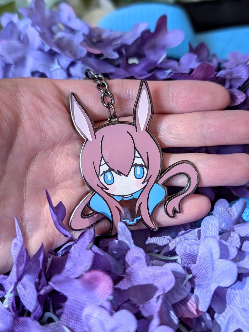 May include: Pink and blue anime-style bunny keychain. The keychain features a cartoon bunny with large blue eyes, pink hair, and a blue collar. The keychain is made of metal and has a black chain. The background is filled with purple flowers.