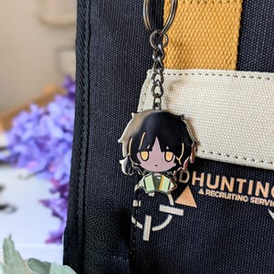 May include: A metal keychain featuring a chibi anime character with black hair and yellow eyes. The keychain is attached to a black and tan tote bag with white text that reads "DHUNTING A RECRUITING SERVICE".