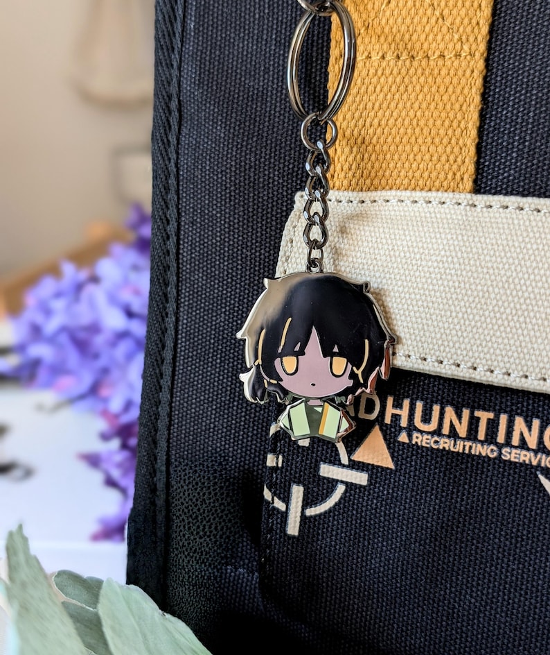 May include: A metal keychain featuring a chibi anime character with black hair and yellow eyes. The keychain is attached to a black and tan tote bag with white text that reads "DHUNTING A RECRUITING SERVICE".