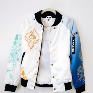 May include: White bomber jacket with a black collar and cuffs. The jacket has gold and blue embroidery on the sleeves and a gold embroidered design on the front. The zipper has a gold pull with the word "YAKSHA" printed on it.