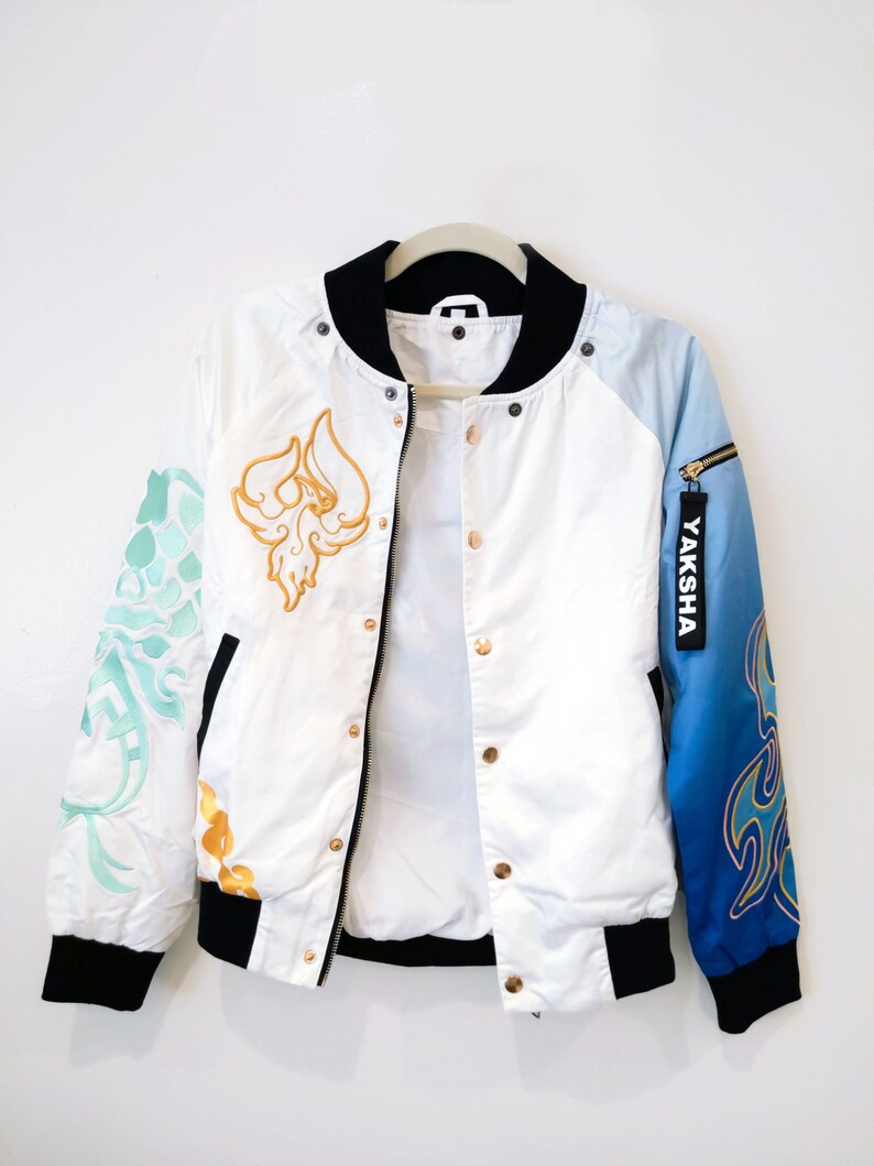May include: White bomber jacket with a black collar and cuffs. The jacket has gold and blue embroidery on the sleeves and a gold embroidered design on the front. The zipper has a gold pull with the word "YAKSHA" printed on it.