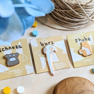 May include: Three enamel pins with different designs. The first pin is a brown dog with the text "Schnozz" above it. The second pin is a light blue elephant with the text "burz" above it. The third pin is an orange fox with the text "Shobe" above it. Each pin has the text "enamel pin" below the design.