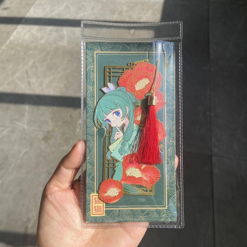 May include: A bookmark featuring an anime-style illustration of a girl with teal hair and blue eyes, wearing a green kimono. The bookmark has a red tassel and is decorated with red flowers. The bookmark is in a clear plastic sleeve. The text "猫猫" is visible.