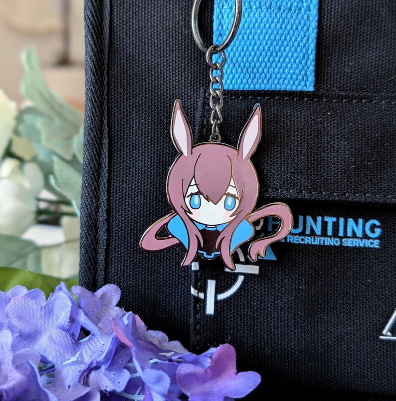 May include: A metal keychain featuring a cartoon character with bunny ears, blue eyes, and a blue and pink outfit. The keychain is attached to a silver chain and ring. The background includes a black bag with the text "HUNTING RECRUITING SERVICE".