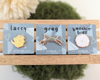 May include: Three enamel pins on display. One is yellow and shaped like a dog with the name "larry". Another is gray and shaped like a dog with the name "greg". The third is white with the text "b*tchin' fries".