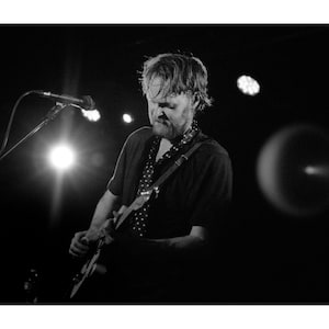 Pode incluir: Uma fotografia a preto e branco de um músico a tocar uma guitarra elétrica num palco. O músico está a usar uma camisa preta com um padrão de bolinhas brancas e tem um microfone à sua frente.