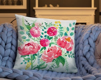 Puede incluir: Una almohada blanca con un diseño de acuarela de peonías rosas y hojas verdes. La almohada está sobre una manta de punto azul.