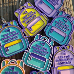 May include: A collection of backpack-shaped keychains in various color combinations including purple, yellow, teal, and light pink. Each keychain features a raised NFC symbol and the embossed text TAP FOR EMERGENCY CONTACT on the front.