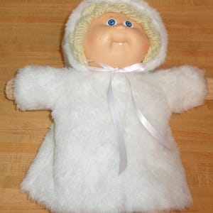 May include: A Cabbage Patch Kid doll wearing a white fur coat with a white satin ribbon tied around the waist. The doll has blonde hair and blue eyes.