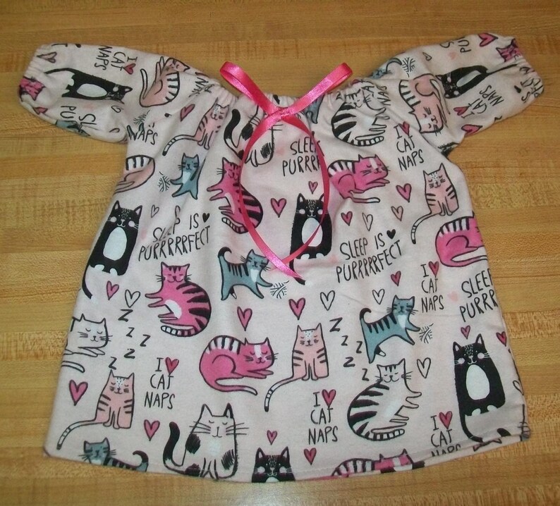 May include: A pale pink nightdress featuring a cat print, short sleeves, and a pink ribbon. The fabric is decorated with cat drawings, hearts, and the phrases "Sleep is Purrrfect" and "I love cat naps."