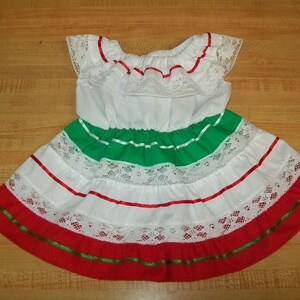 Puede incluir: Un vestido blanco con ribetes de cinta roja, verde y blanca. El vestido tiene un escote de encaje y una falda con volantes.