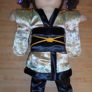 Peut inclure: Une poupée vêtue d'un mini-kimono. Le kimono crème présente un motif floral et une bordure noire. La tenue comprend une ceinture obi noire avec un cordon doré et un pantalon noir. La poupée a des couettes brunes avec des rubans violets.
