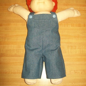 May include: A vintage doll with a vinyl face, orange yarn hair, and blue denim overalls. The doll has its arms outstretched and is lying on a wood-grain surface. The overalls have two blue buttons on the straps and a front pocket.