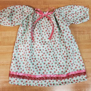 Pode incluir: Um vestido azul claro com um padrão floral de pequenas flores vermelhas. O vestido tem mangas curtas bufantes e um laço rosa no decote. Uma fita rosa e renda adornam a bainha.