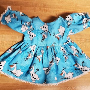May include: A light blue baby dress featuring a print of the snowman Olaf from the movie Frozen. The dress has short, puffy sleeves and a flared skirt, trimmed with white lace. The Olaf print is repeated across the fabric.