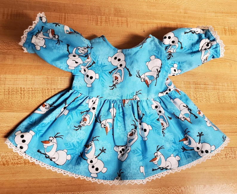 May include: A light blue baby dress featuring a print of the snowman Olaf from the movie Frozen. The dress has short, puffy sleeves and a flared skirt, trimmed with white lace. The Olaf print is repeated across the fabric.
