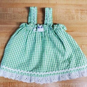 May include: A green and white gingham dress with a white lace trim and a cat button. The dress has thin straps and a gathered bodice.