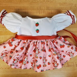Op de afbeelding: Een witte top met pofmouwen en rode afwerking, gecombineerd met een roze rok met een aardbeienkarakterprint. De outfit heeft een rode satijnen lintriem en aardbeivormige knopen.