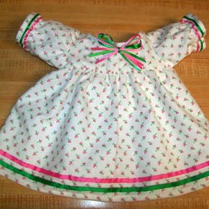 May include: A white baby dress with a floral pattern of small pink flowers and green leaves. The dress has green and pink ribbon accents on the sleeves and hem, and a bow at the neckline. The dress has short sleeves.