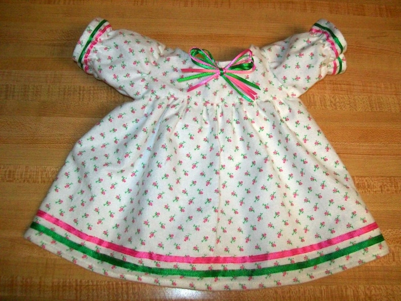 May include: A white baby dress with a floral pattern of small pink flowers and green leaves. The dress has green and pink ribbon accents on the sleeves and hem, and a bow at the neckline. The dress has short sleeves.