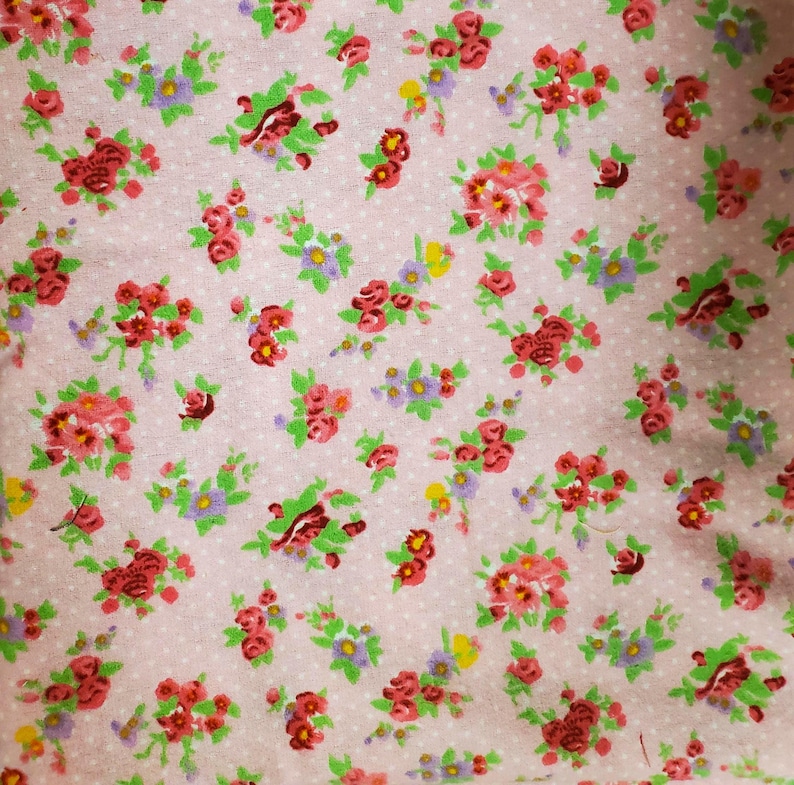 May include: A close-up of a pink fabric with a floral pattern. The pattern features clusters of red, pink, purple, and yellow flowers with green leaves. The background is a light pink with small white polka dots.