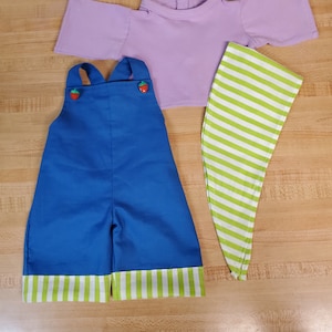 Peut inclure: Une tenue de poupée avec une chemise lavande, une salopette bleue à rayures vertes et blanches et un chapeau rayé vert et blanc.